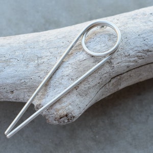 Pode incluir: Um grampo de cabelo prateado com um design simples e curvo. O grampo é feito de uma única peça de metal e tem um acabamento liso e polido.