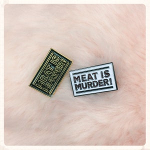 Puede incluir: Dos pines de esmalte con el texto "Meat is Murder!" en letras blancas y negras. Los pines están sobre un fondo rosa difuso.