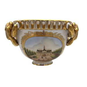 Puede incluir: Un tazón de porcelana blanca con ribetes dorados y dos asas doradas ornamentadas. El tazón presenta una escena pintada de un edificio con un cielo azul y árboles verdes.