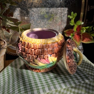 May include: A brown and green ceramic sugar bowl with a floral design and a separate lid. The bowl has a textured surface and a purple interior.