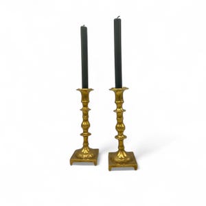 Peut inclure: Une paire de bougeoirs en métal doré avec des bougies noires. Les bougeoirs ont une base carrée et un design cannelé.