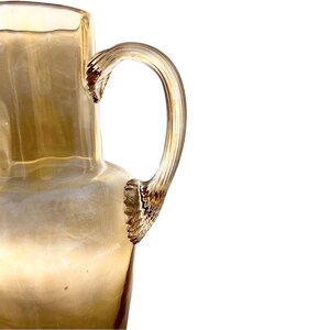 Op de afbeelding: Een heldere glazen kan met een getextureerde amberkleurige handgreep. De kan heeft een rond lichaam en een brede opening.