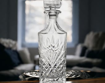 May include: A clear glass decanter with a square stopper. The decanter has a rectangular body with a detailed, cut-glass pattern. The stopper is also square and clear. The design is suitable for spirits or wine.
