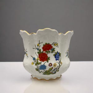 Puede incluir: Un macetero de porcelana blanca con un borde festoneado y un ribete dorado. El macetero está decorado con un patrón floral en tonos rojos, azules, amarillos y verdes.
