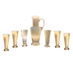 Op de afbeelding: Een set van zeven heldere glazen drinkglazen, inclusief een kan. De glazen zijn allemaal van dezelfde vorm en grootte, met een uitlopende bovenkant en een ronde basis. De kan heeft een handvat en een tuit. De glazen en de kan zijn allemaal licht amberkleurig.