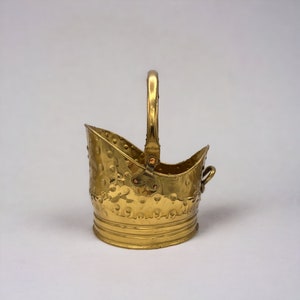 Puede incluir: Un pequeño cubo de carbón de metal dorado con un acabado martillado y un asa. El cubo tiene un fondo redondeado y una parte superior ligeramente acampanada.