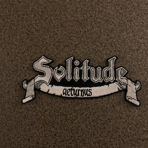 May include: A white embroidered patch with the words "Solitude Aeturnus" in a black banner.