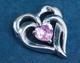 Peut inclure: Pendentif argenté en forme de cœur avec un petit cœur rose en son centre. Le pendentif a une finition polie et se détache sur un fond bleu foncé. La pierre précieuse est de couleur rose clair.