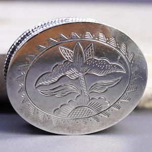 May include: An oval-shaped silver-toned metal box featuring an engraved floral design on the lid. The center of the lid displays a stylized flower with petals and leaves, surrounded by a decorative border of repeating triangular hatch marks.