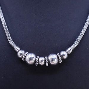Puede incluir: Un collar de plata con una serie de cuentas redondas y pulidas de diferentes tamaños. Las cuentas están ensartadas en una cadena trenzada. El collar se muestra sobre un fondo oscuro.