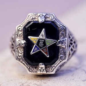 May include: A silver ring featuring an octagonal black stone. A multi-coloured star emblem is set in the centre of the black stone. The ring has an ornate, filigree design on the band and around the stone.