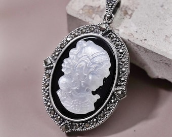 May include: Oval pendant with a cameo design. The pendant features a white profile against a black background, framed by a silver-colored border with small, decorative accents. The pendant has a loop at the top for a chain.