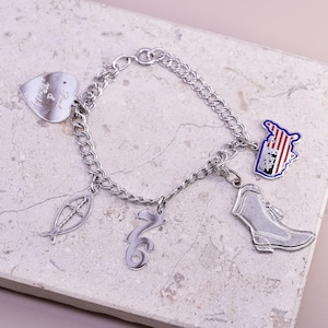 May include: Silver charm bracelet with various charms, including a heart with text, a fish symbol, a stylized number, a boot, and a patriotic emblem with red and blue accents. The bracelet is on a light-colored surface.