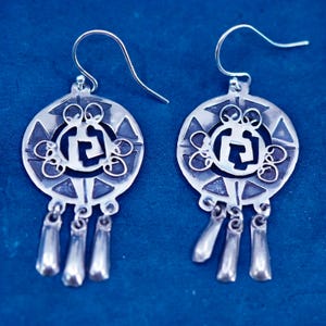 Puede incluir: Un par de pendientes plateados. Cada pendiente presenta un diseño circular con una forma geométrica central, rodeada de elementos decorativos. Tres colgantes en forma de lágrima cuelgan de la parte inferior de cada pendiente. Cierre de gancho.