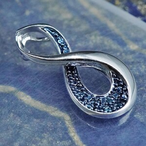 May include: A silver-toned infinity pendant with a polished finish. The pendant features a design filled with small, dark blue gemstones. The infinity symbol is a classic jewelry design, often symbolizing eternity or endless love.