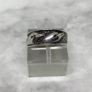 Op de afbeelding: Een zilveren ring met een herhaald patroon van gestileerde vogelkoppen. De ring heeft een getextureerde band en wordt gepresenteerd op een transparante standaard tegen een zachte, neutrale achtergrond. Het ontwerp van de ring is gedetailleerd en symmetrisch.