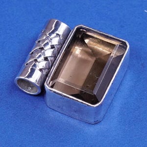 May include: A silver-toned rectangular jewelry box with a large, faceted, smoky quartz gemstone. Beside it is a cylindrical silver piece with a woven design. The items are displayed on a blue surface.