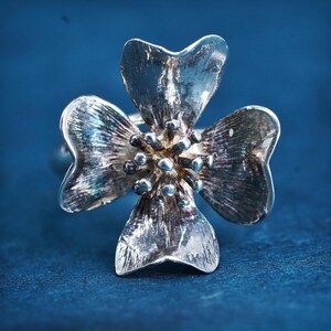 May include: A silver-toned ring featuring a four-petal flower design. The petals have a textured appearance, and the center of the flower is adorned with small, spherical details. The ring is set against a deep blue background.