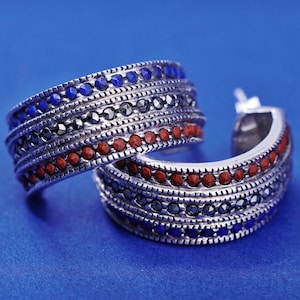 May include: A pair of silver hoop earrings with intricate detailing. The earrings feature alternating rows of blue, black, and red gemstones. The design is set against a bright blue background, highlighting the jewelry's colors and textures.