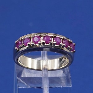 May include: A gold and silver ring with seven round, vibrant red gemstones. The ring is set against a blue background, showcasing the gemstones' color and the ring's design. A piece of jewelry.
