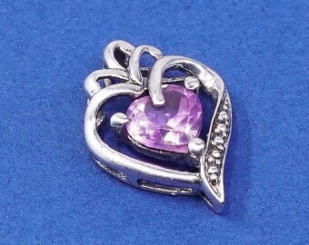 Peut inclure: Pendentif argenté avec une pierre précieuse en forme de cœur, de couleur violet clair. Le pendentif présente un motif ajouré et une rangée de petites pierres claires. Le fond est bleu uni.
