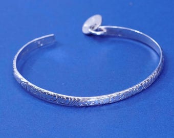 Pode incluir: Uma pulseira de punho prateada com um padrão floral gravado. A pulseira aberta tem uma forma arredondada e um pequeno fecho decorativo. A pulseira é exibida em uma superfície azul.