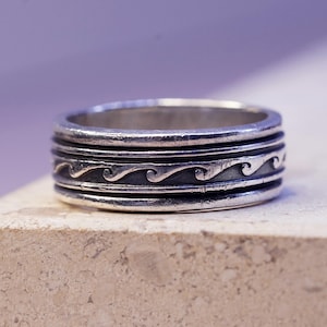 May include: A silver-toned ring with a wave pattern. The ring has a textured surface and a band with a series of horizontal lines. The wave pattern is etched into the band, adding a decorative element. The ring is likely made of metal.