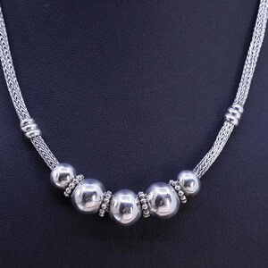 May include: A silver necklace featuring a series of large, polished spheres interspersed with smaller beaded accents. The necklace has a woven chain design on each side, with a clasp at the back. The necklace is displayed against a dark background.