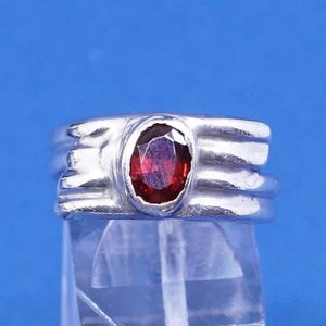 May include: A silver ring with a large, oval-shaped, deep red gemstone. The ring has a textured band with horizontal ridges. The gemstone is set in a bezel setting, and the ring is displayed against a blue background.