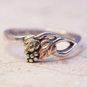May include: Silver ring with a grape and leaf design. The leaves are gold and rose-gold colored, and the grapes are small and silver. The ring has a delicate, nature-inspired aesthetic.