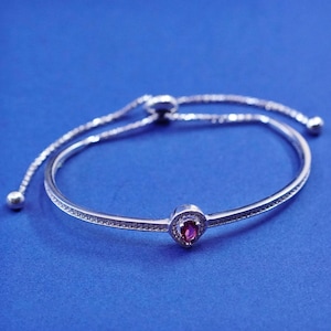 Pode incluir: Uma pulseira prateada com uma pedra central ovalada de cor rubi. A pulseira apresenta um design delicado com pequenas pedras transparentes e um fecho deslizante ajustável. O fundo é azul liso.