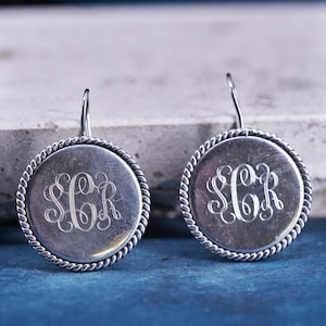 May include: A pair of silver-toned round earrings. Each earring features a monogram in a script font within a circular frame with a twisted rope design. The earrings have a simple hook closure.