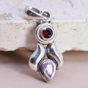 May include: A silver pendant with a red gemstone at the top and a teardrop-shaped purple gemstone at the bottom. The pendant has a unique, flowing design and a loop for a chain.