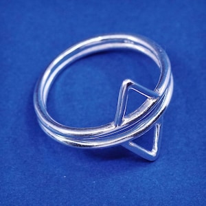 May include: A silver ring with a geometric design. The ring features a double band with a triangular accent. The ring is set against a blue background. The ring is made of a shiny, reflective metal.