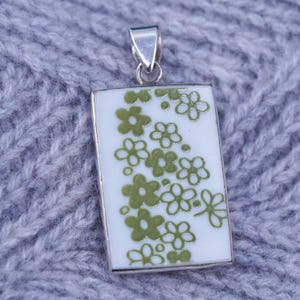 Könnte beinhalten: Rechteckiger Anhänger mit silberner Öse. Der Anhänger hat einen weißen Hintergrund mit einem Muster aus grünen floralen Designs. Der Anhänger ist wahrscheinlich aus Keramik oder Emaille.
