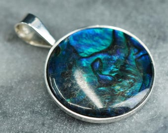 May include: A round pendant with a vibrant, iridescent abalone shell center, showcasing shades of blue and green. The shell is set in a silver-colored metal frame with a matching bail for a necklace chain.