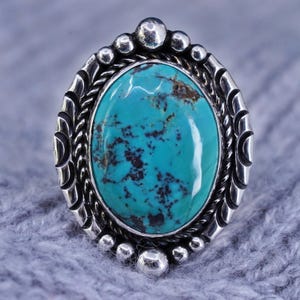 Pode incluir: Anel oval de turquesa com moldura de prata. A turquesa é de cor azul-esverdeada vibrante com padrões castanhos escuros. A moldura de prata tem um design detalhado com detalhes de contas e corda.