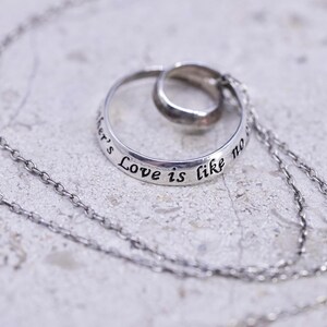 May include: A silver necklace featuring a pendant with two interlocking rings. The larger ring is inscribed with the words "Love is like no other's". The chain is delicate and the pendant is centered. The background is a light gray surface.