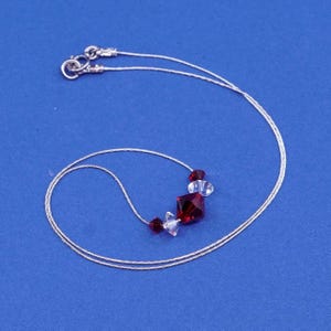 May include: A delicate silver necklace with a pendant featuring a large, faceted, square, deep red crystal, flanked by smaller red and clear crystals. The necklace has a simple, elegant design, perfect for adding a touch of sparkle.