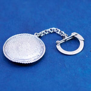 May include: A silver-toned round pendant with an engraved Aztec calendar design, connected by a chain to a clasp. The pendant has a detailed pattern and is set against a blue background. The clasp is a simple, curved design.