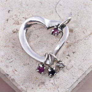 May include: A silver-toned heart-shaped pendant with three small charms and two gemstones. The pendant has a loop at the top for a chain. The charms include a ruby-colored gemstone, a dark blue gemstone, and a clear gemstone.