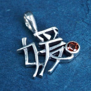 May include: A silver pendant with Chinese characters and a small, round, red gemstone. The pendant has a loop at the top for a chain. The background is a dark blue.