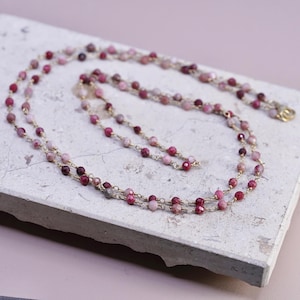 May include: A delicate necklace featuring small, round, pink and burgundy beads strung on a gold-colored chain. The necklace is displayed on a light gray surface, showcasing its layered design and intricate details. The beads vary in shade, creating a gradient effect.