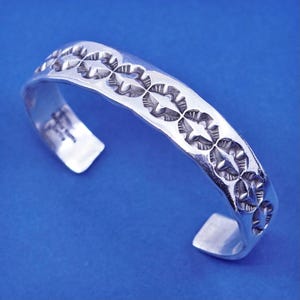 May include: A silver cuff bracelet with a repeating star and leaf pattern. The open-ended bracelet has a slightly hammered texture and is set against a blue background. The bracelet is a piece of jewelry.