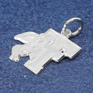 May include: A silver pendant featuring a cross design with an angel figure. The pendant has a detailed, three-dimensional appearance and includes a small loop for attaching to a chain. The pendant is displayed against a textured blue background.