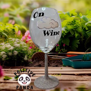 May include: A clear wine glass with a silver glitter stem. The glass has the words "On Wine" with a cloud graphic. The background is a blurred garden scene with wooden planks.