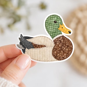 May include: A sticker of a duck with a patchwork design. The duck has a green checkered head, a yellow beak, and a brown floral patterned body. The wings are a mix of beige, brown, and gray fabrics. The sticker is held by a person.