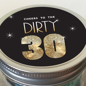 Op de afbeelding: Zwarte potdeksel met een gouden glitter "Dirty 30" ontwerp en de tekst "Cheers to the" boven het nummer. De deksel heeft een zilveren rand.