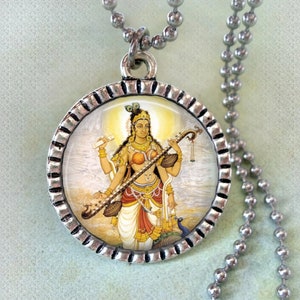 Puede incluir: Un collar de plata con un colgante redondo que presenta una ilustración colorida de una diosa hindú tocando un instrumento de cuerda. El colgante tiene un borde de plata con un diseño texturizado.