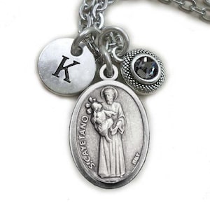 Op de afbeelding: Een zilverkleurige ketting met een hanger met een afbeelding van een heilige, een ronde bedel met de letter "K" en een kleine, donkergrijze edelsteen. De ketting heeft een schakelkettingontwerp. De achtergrond is wit.